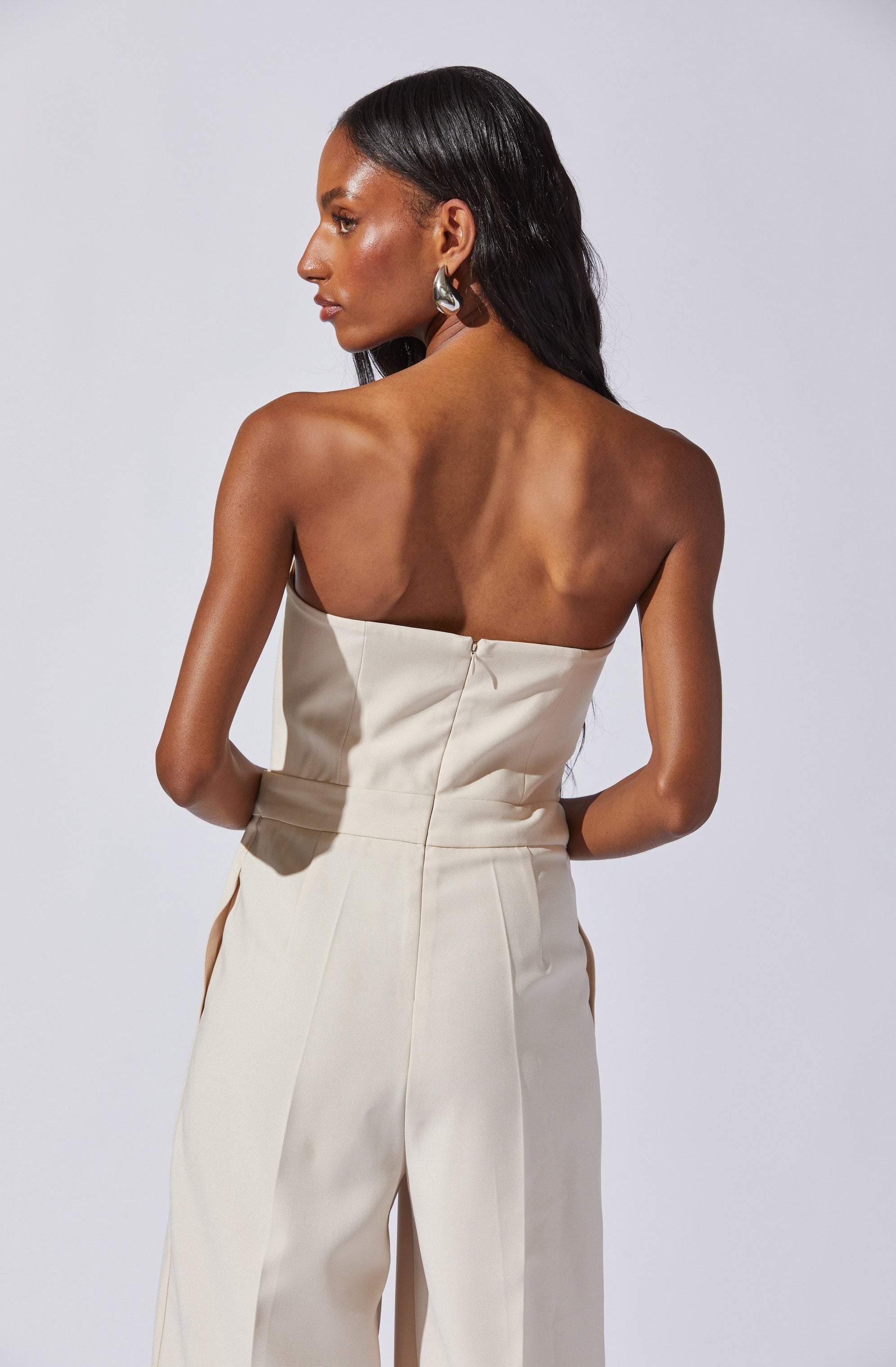 A woman is wearing a strapless, cream-colored jumpsuit with a fitted bodice and wide-leg pants. She has long, straight black hair and is accessorized with large, silver hoop earrings.