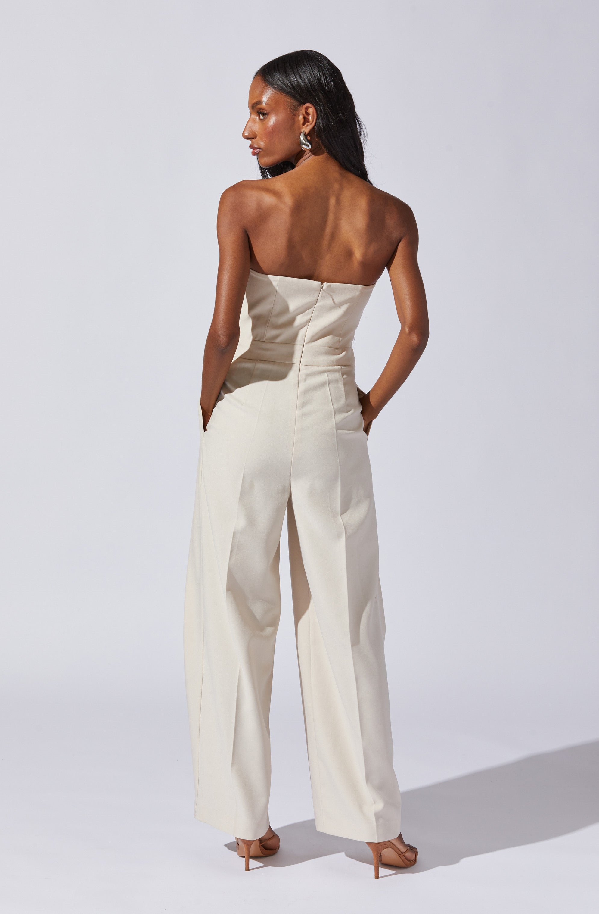 A woman is wearing a strapless, cream-colored jumpsuit with wide-leg pants. She has her hands in her pockets and is accessorized with large, silver earrings. She is also wearing open-toed, nude-colored high heels.