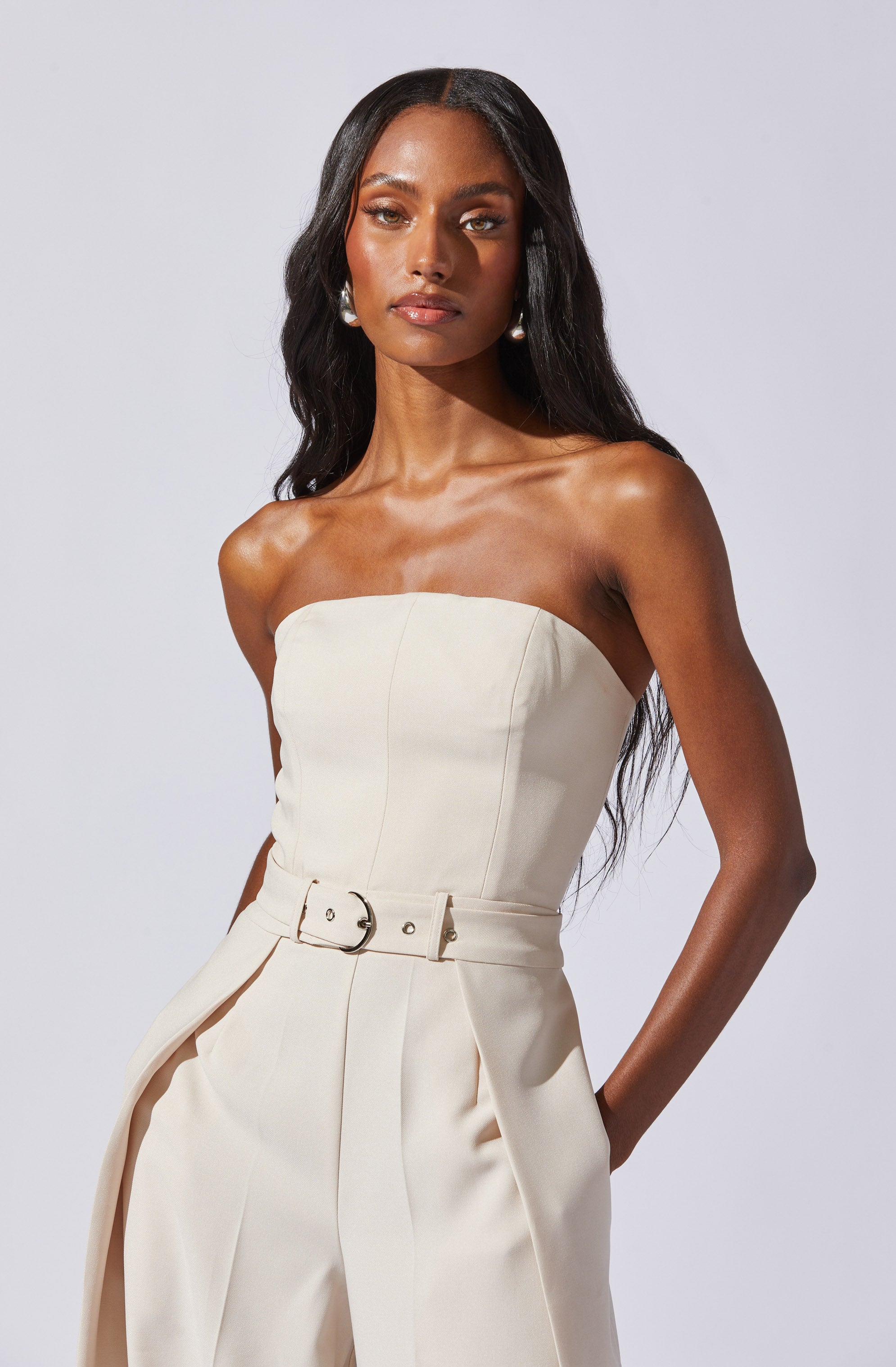 A woman is wearing a strapless, off-white jumpsuit with a fitted bodice featuring vertical seams. The jumpsuit has a matching belt with a round buckle at the waist, and the pants are high-waisted with pleats. The fabric appears smooth and structured.