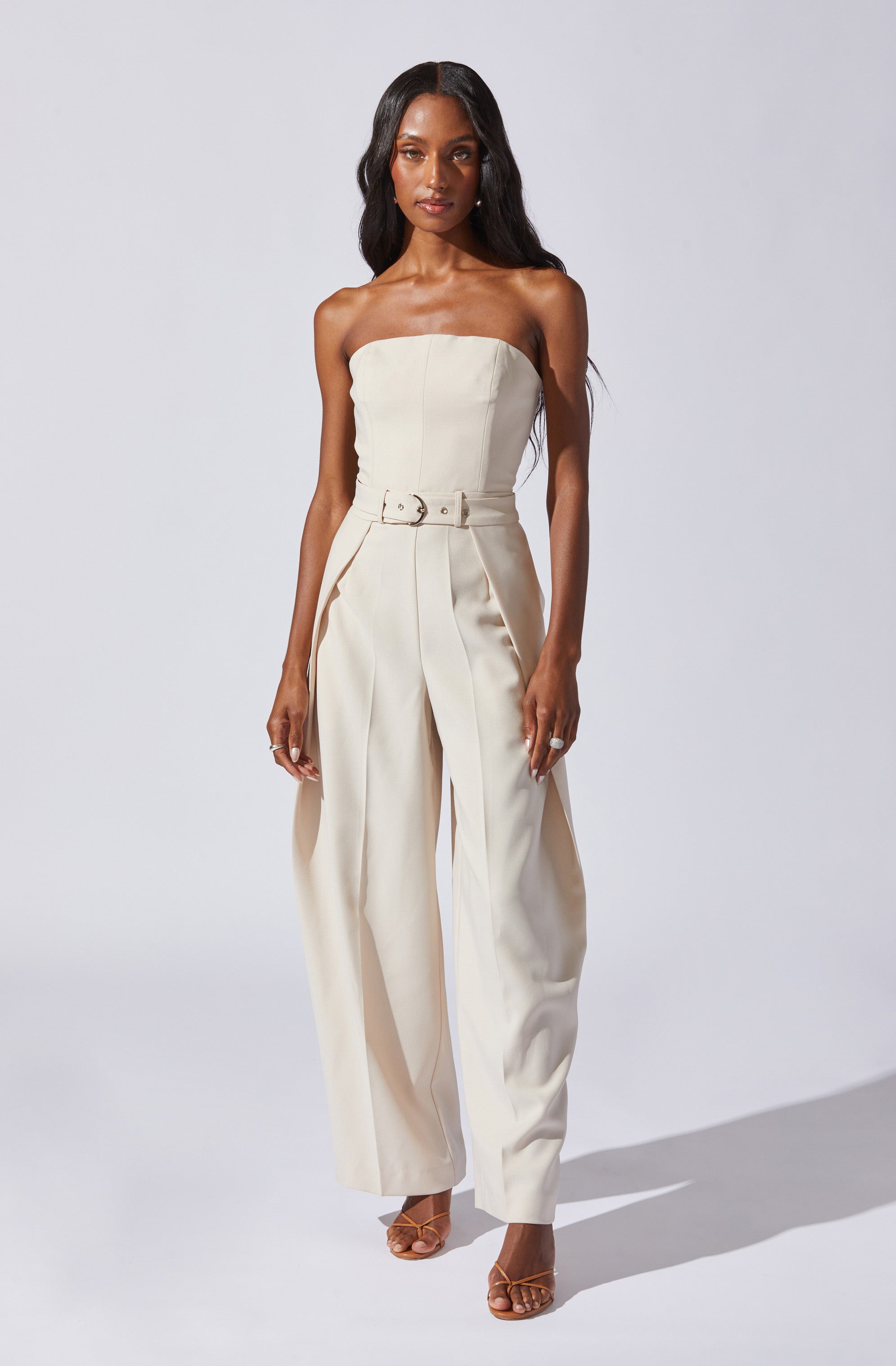 A woman is wearing a strapless, off-white jumpsuit with a fitted bodice and wide-leg trousers. The jumpsuit features a matching belt at the waist with a round buckle. She is also wearing open-toed sandals with thin straps.