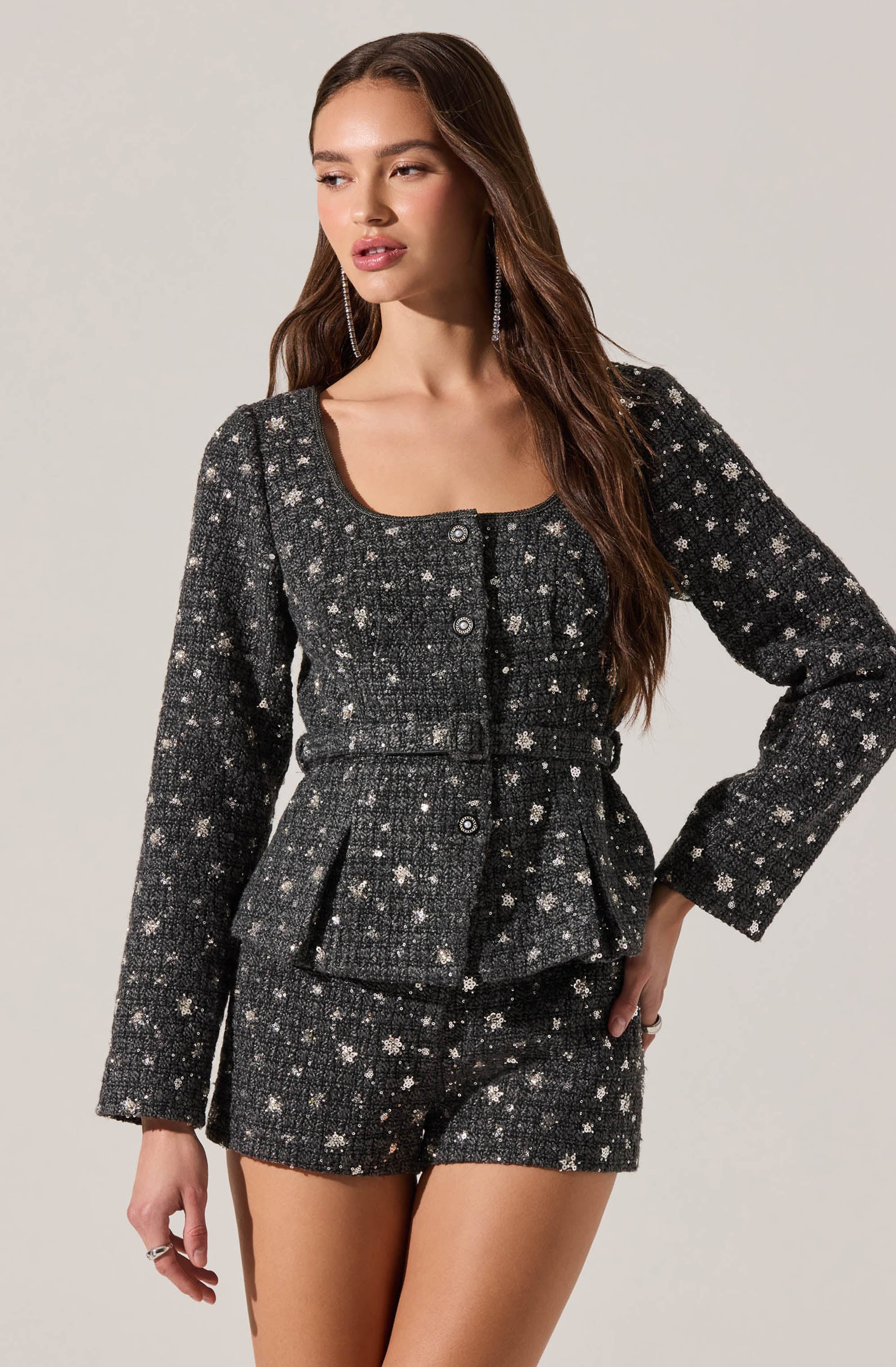 A woman is wearing a matching black tweed suit with a textured pattern of white and silver stars. The suit includes a fitted, long-sleeved jacket with a rounded neckline, button-front closure, and two flap pockets at the waist. She is also wearing high-waisted shorts made from the same star-patterned tweed fabric.