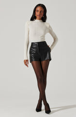 A woman is wearing a long-sleeve, cream-colored, ribbed sweater with a high neckline. She pairs it with high-waisted black leather shorts and sheer black tights. She completes her outfit with black pointed-toe heels.