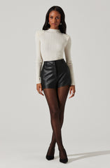 A woman is wearing a long-sleeved, cream-colored ribbed sweater with a high neckline. She pairs it with high-waisted, black leather shorts. She is also wearing sheer black tights and black pointed-toe heels. Her accessories include earrings and rings.