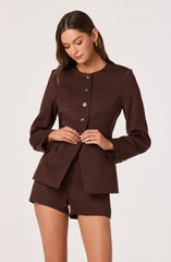 A woman is wearing a dark brown tailored blazer with a round neckline and a row of buttons down the front. The blazer has two flap pockets at the waist and buttoned cuffs on the sleeves. She pairs it with matching dark brown shorts. She is also wearing gold hoop earrings and is holding her hands near her waist.
