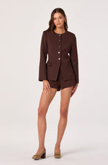 A woman is wearing a dark brown tailored blazer with a round neckline and long sleeves. The blazer features six buttons running down the front and has two flap pockets on either side at the waist. She pairs the blazer with matching dark brown shorts and beige high-heeled shoes with a decorative bow detail on the front. She has medium-length, wavy brown hair and is wearing gold hoop earrings.