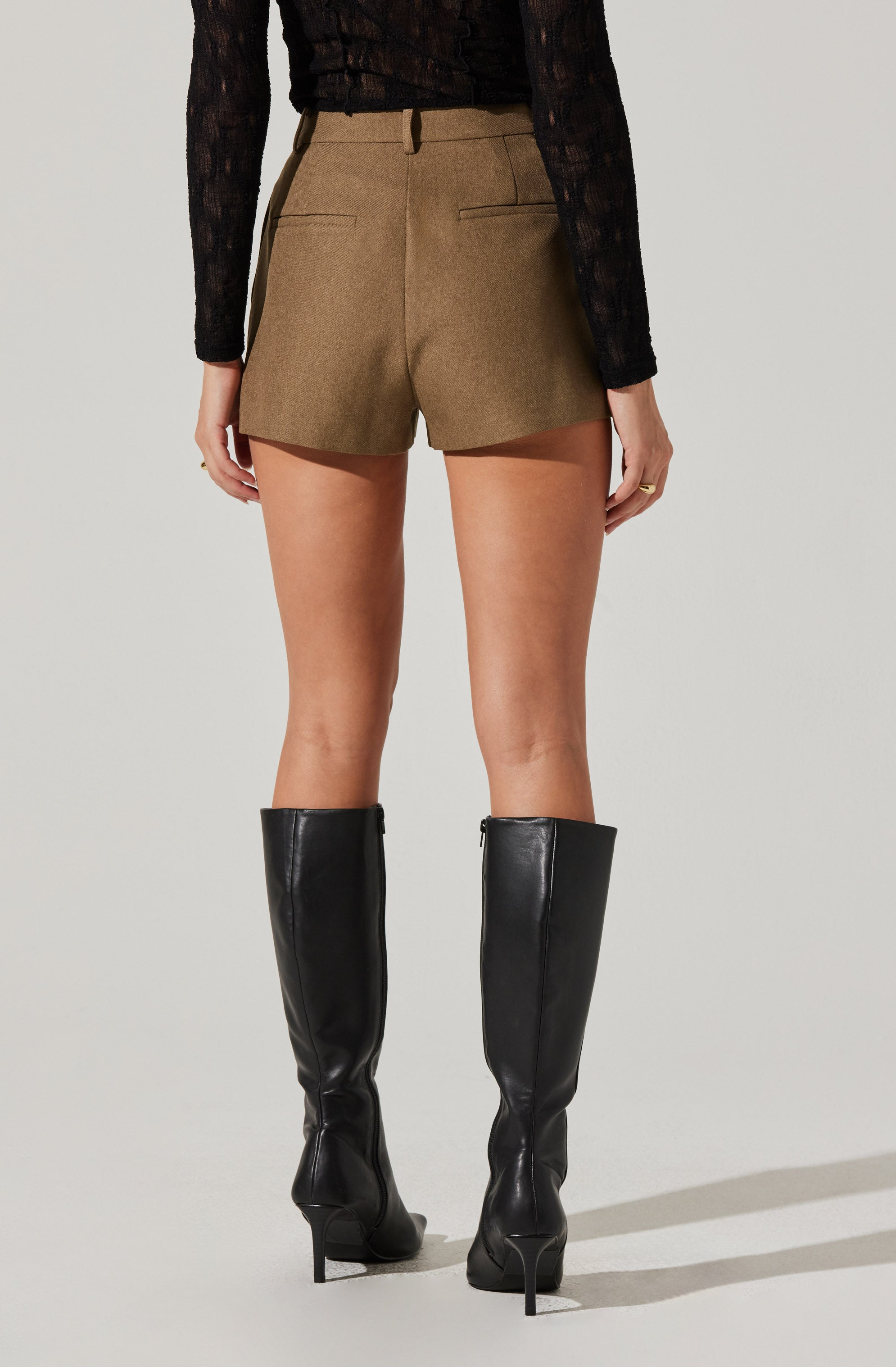 The image shows a person wearing high-waisted brown shorts with a tailored fit, featuring back welt pockets and belt loops. They are also wearing a long-sleeved black lace top, and knee-high black leather boots with stiletto heels.
