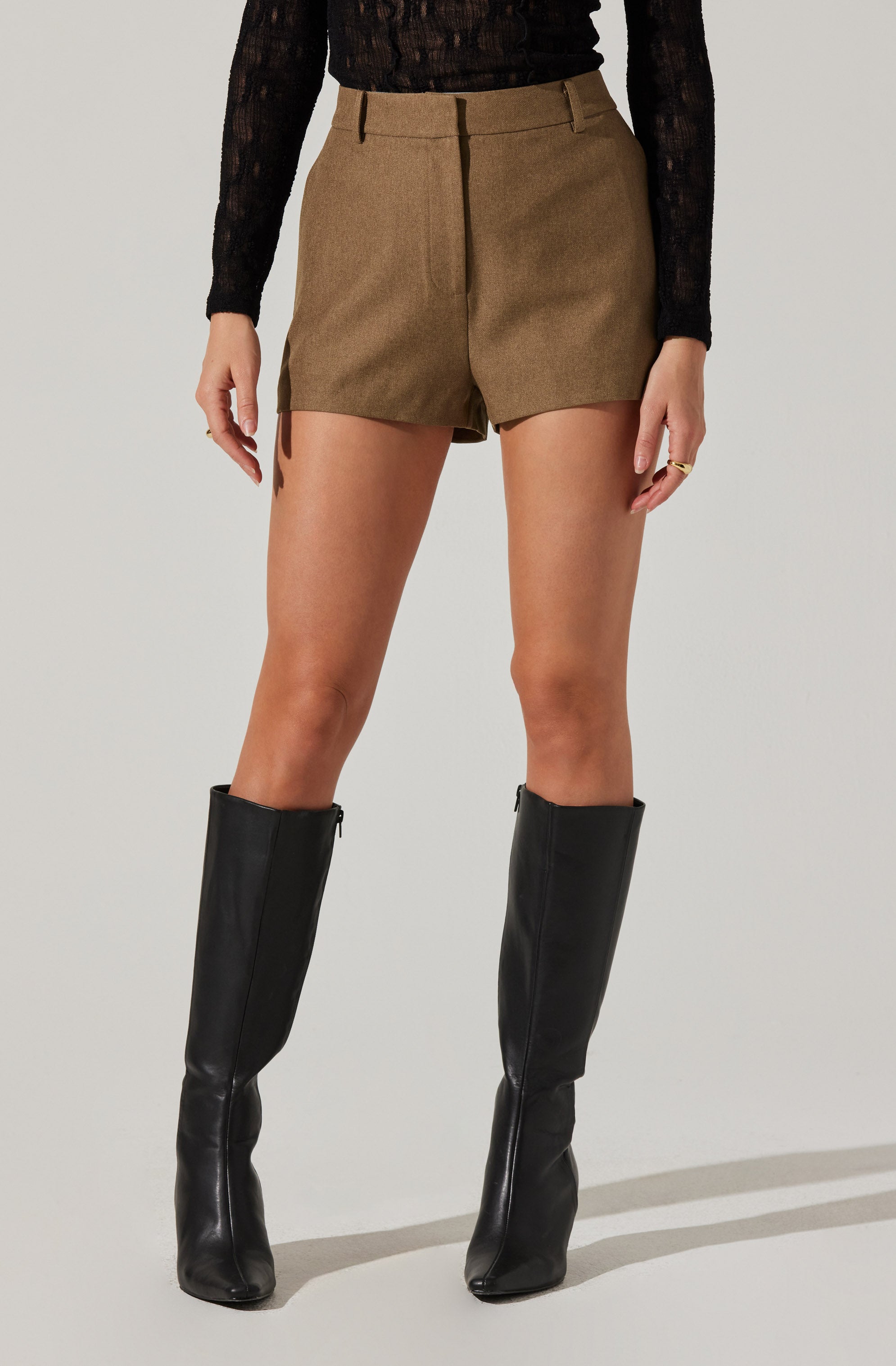 The person is wearing a pair of tailored, high-waisted shorts in a brown or khaki color with belt loops. They are also wearing tall black leather boots that reach just below the knees. The outfit includes a long-sleeved, black, semi-sheer top with a subtle pattern.