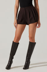 The person is wearing high-waisted dark brown pleated shorts with a subtle pinstripe pattern, paired with a fitted black sleeveless top. They have on tall black leather knee-high boots with a front zipper.