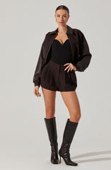 A woman is wearing a black strapless top with a sweetheart neckline, high-waisted dark brown shorts with a textured pattern, a dark brown and black jacket with a zippered front, and knee-high shiny black leather boots with stiletto heels.