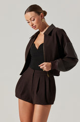 A woman is wearing a black fitted top with a V-neckline, paired with a dark brown, high-waisted pleated skirt that has a zipper detail on the side. She also has on a dark brown bomber jacket with a ribbed collar and cuffs, and leather-like material on the sleeves.