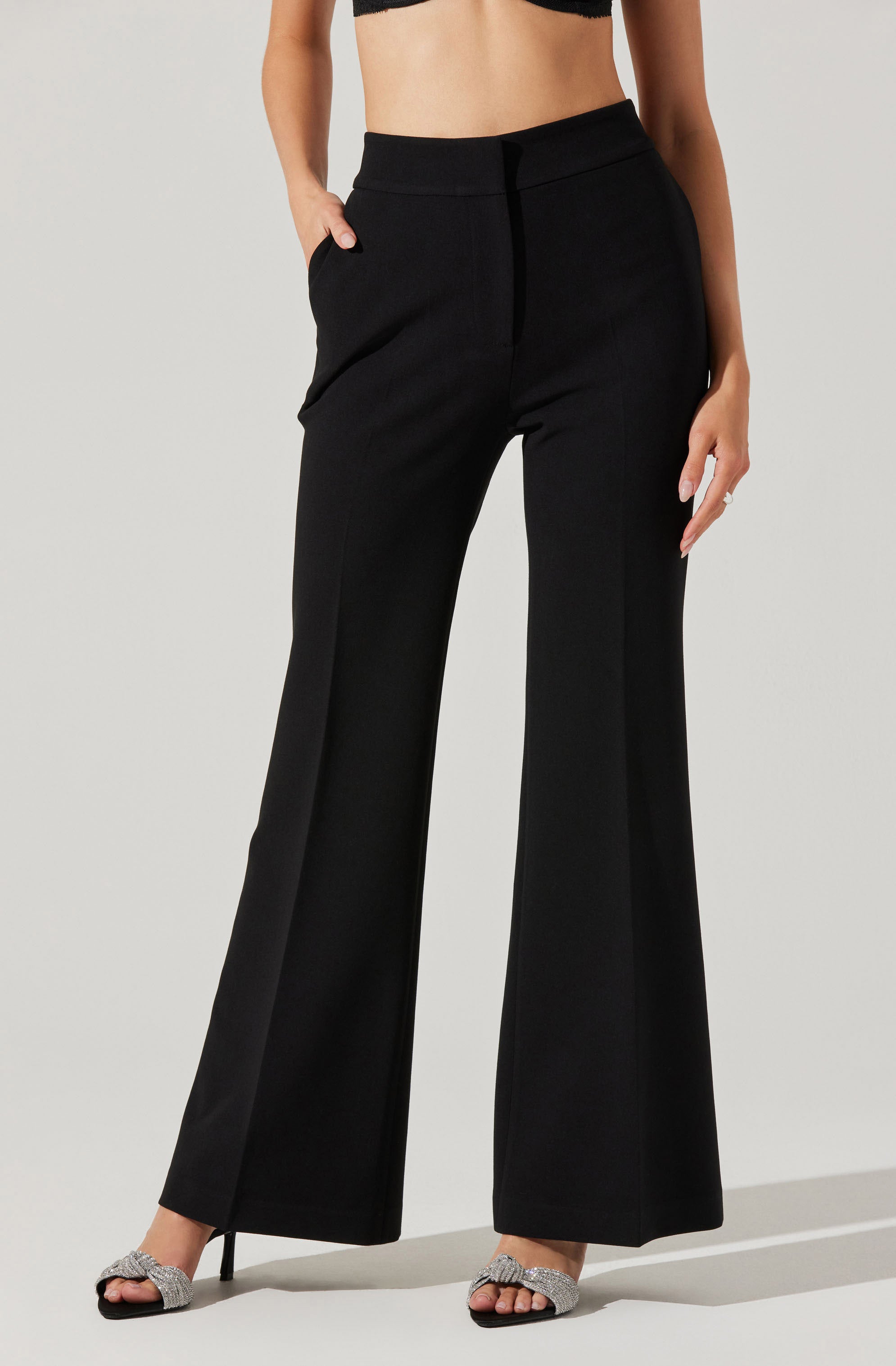 A woman is wearing high-waisted black wide-leg pants with a sleek, tailored appearance. The pants have a smooth waistband without visible belt loops and feature side pockets. She is also wearing a black cropped top, revealing her midriff, and silver high-heeled sandals with a sparkly finish.