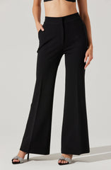 A woman is wearing high-waisted black wide-leg pants with a sleek, tailored appearance. The pants have a smooth waistband without visible belt loops and feature side pockets. She is also wearing a black cropped top, revealing her midriff, and silver high-heeled sandals with a sparkly finish.