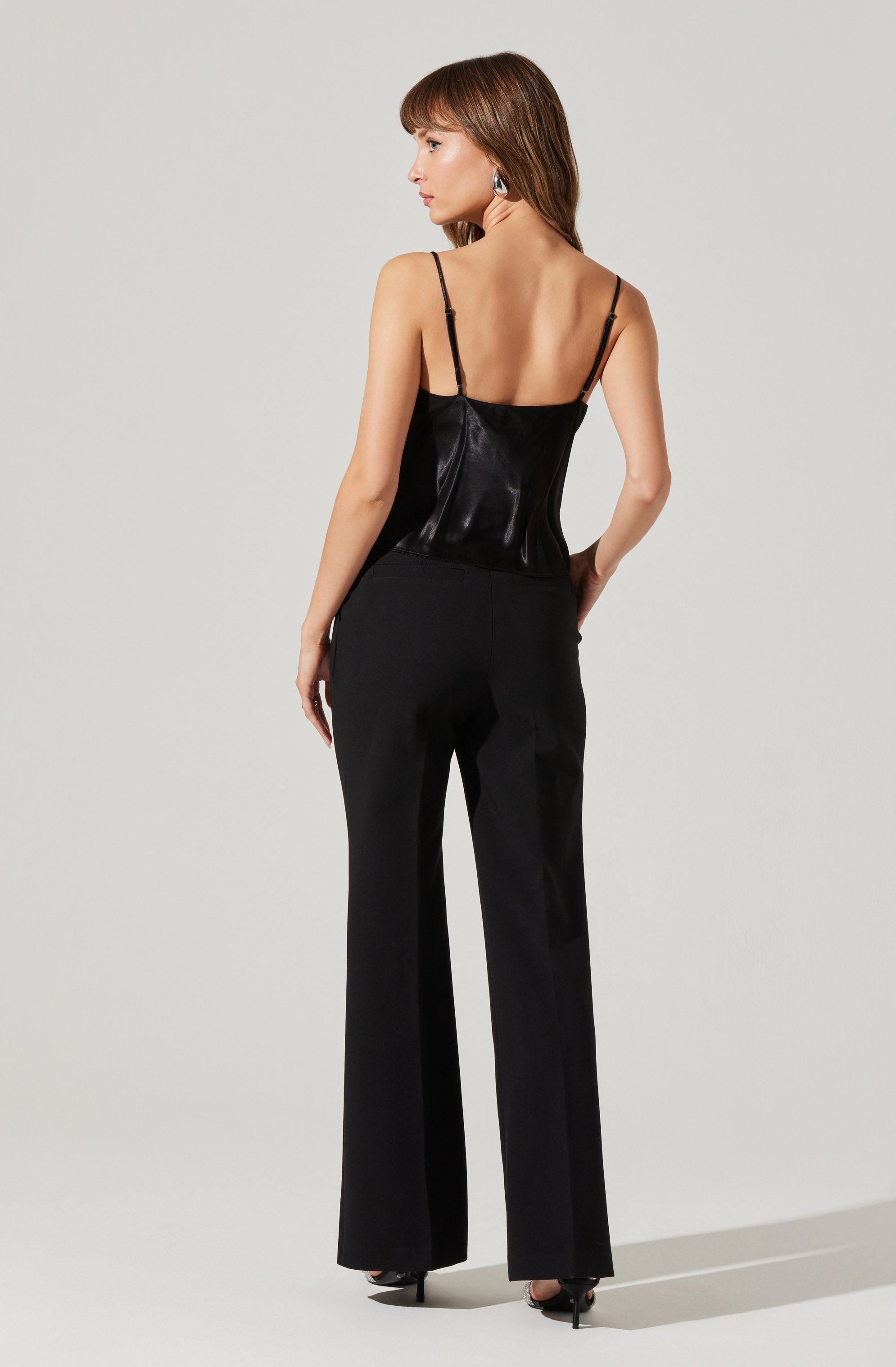 A woman is wearing a black satin camisole top with thin spaghetti straps and a pair of high-waisted, wide-leg black trousers.