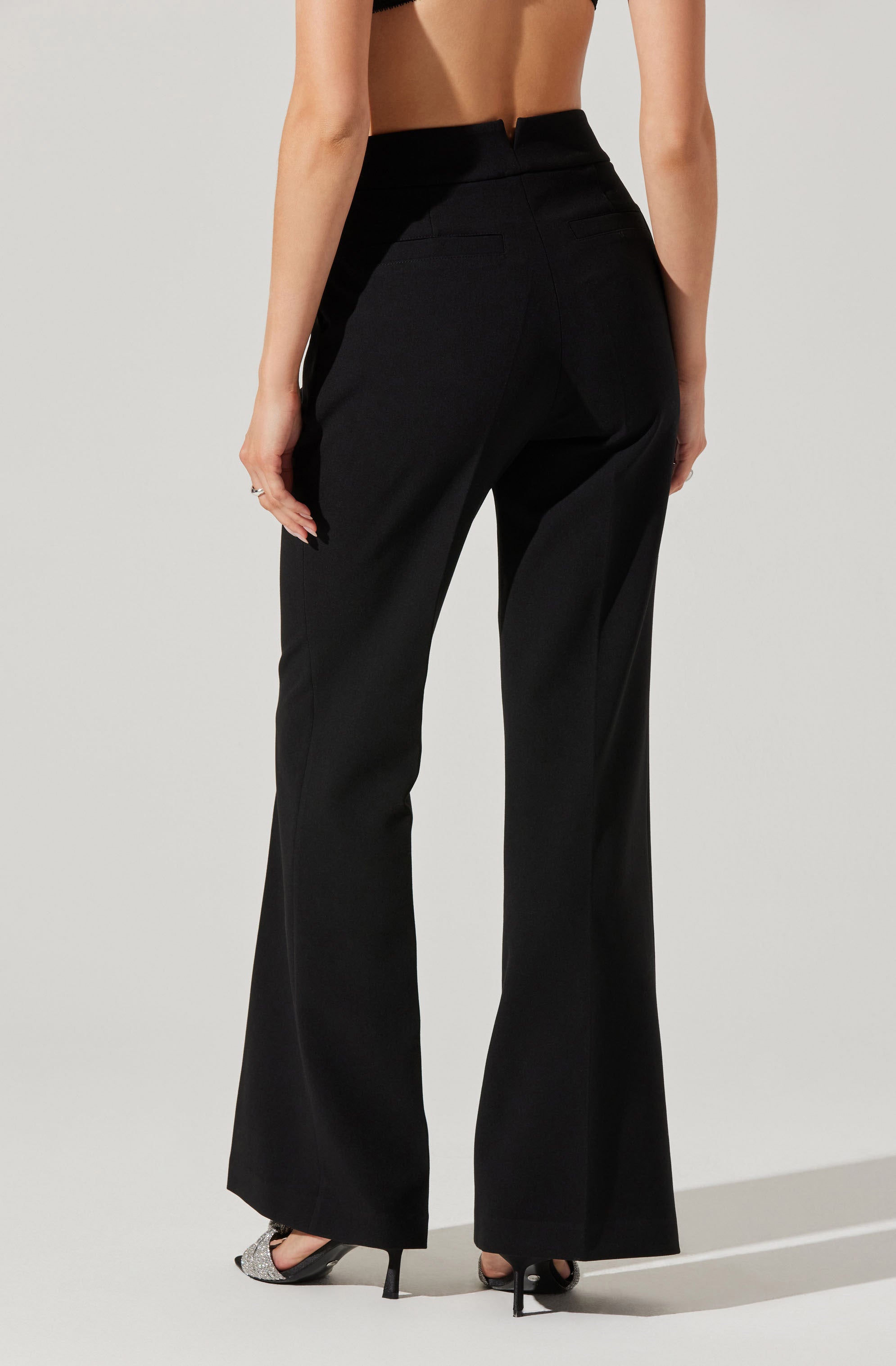 A person is wearing high-waisted black pants with a wide-leg fit. The pants have a structured waistband and appear tailored, with a smooth, solid black fabric. The individual is also wearing black open-toe high heels with a decorative element on the toe strap.