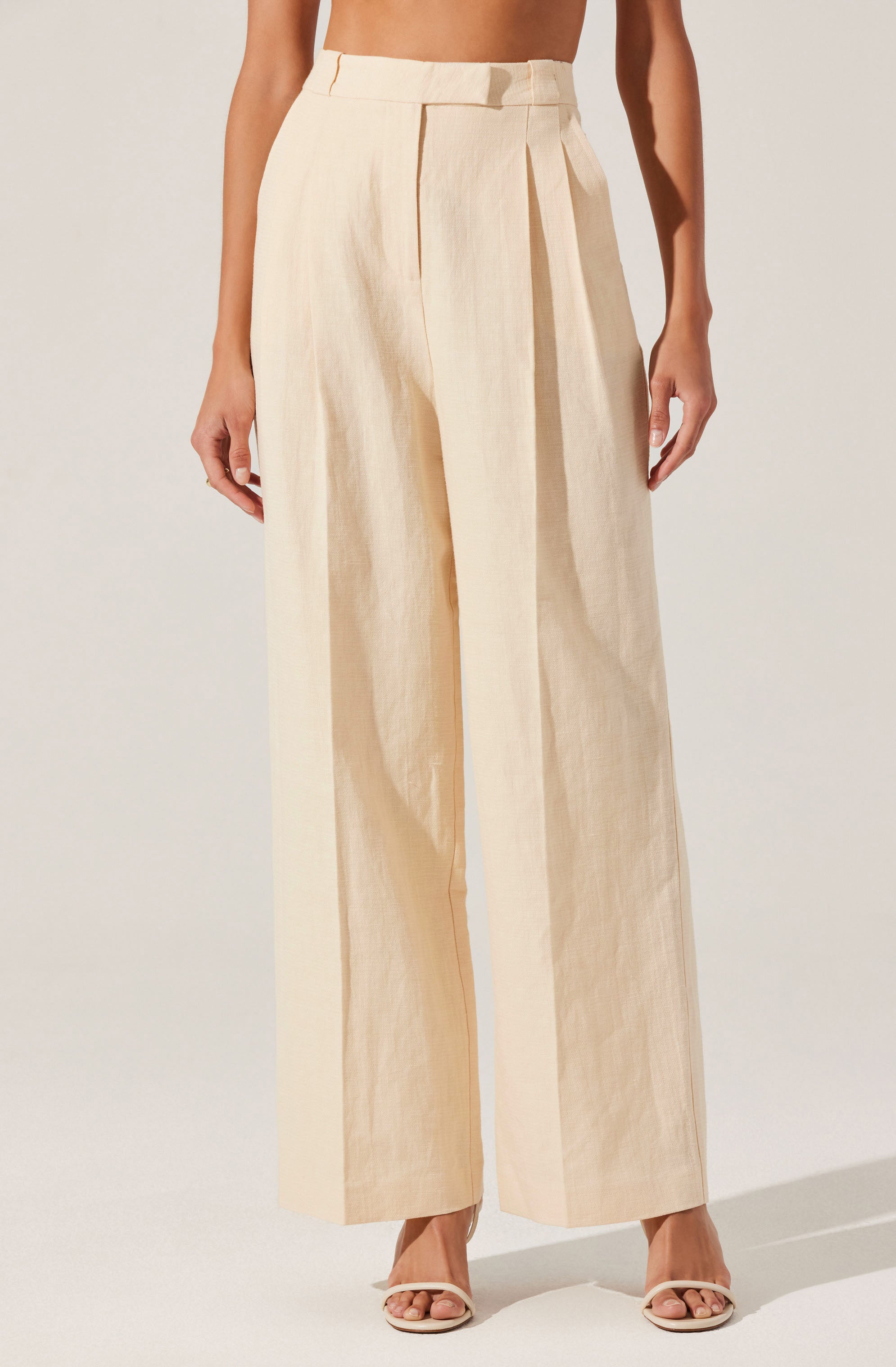 A person is wearing high-waisted, wide-leg trousers in a light beige or cream color. The trousers feature a belt loop detail at the waistband and a prominent front pleat that runs down each leg. The fabric appears to be lightweight and slightly textured. The outfit is paired with open-toed heels. The person's arms are relaxed by their sides, and the background is plain and neutral.