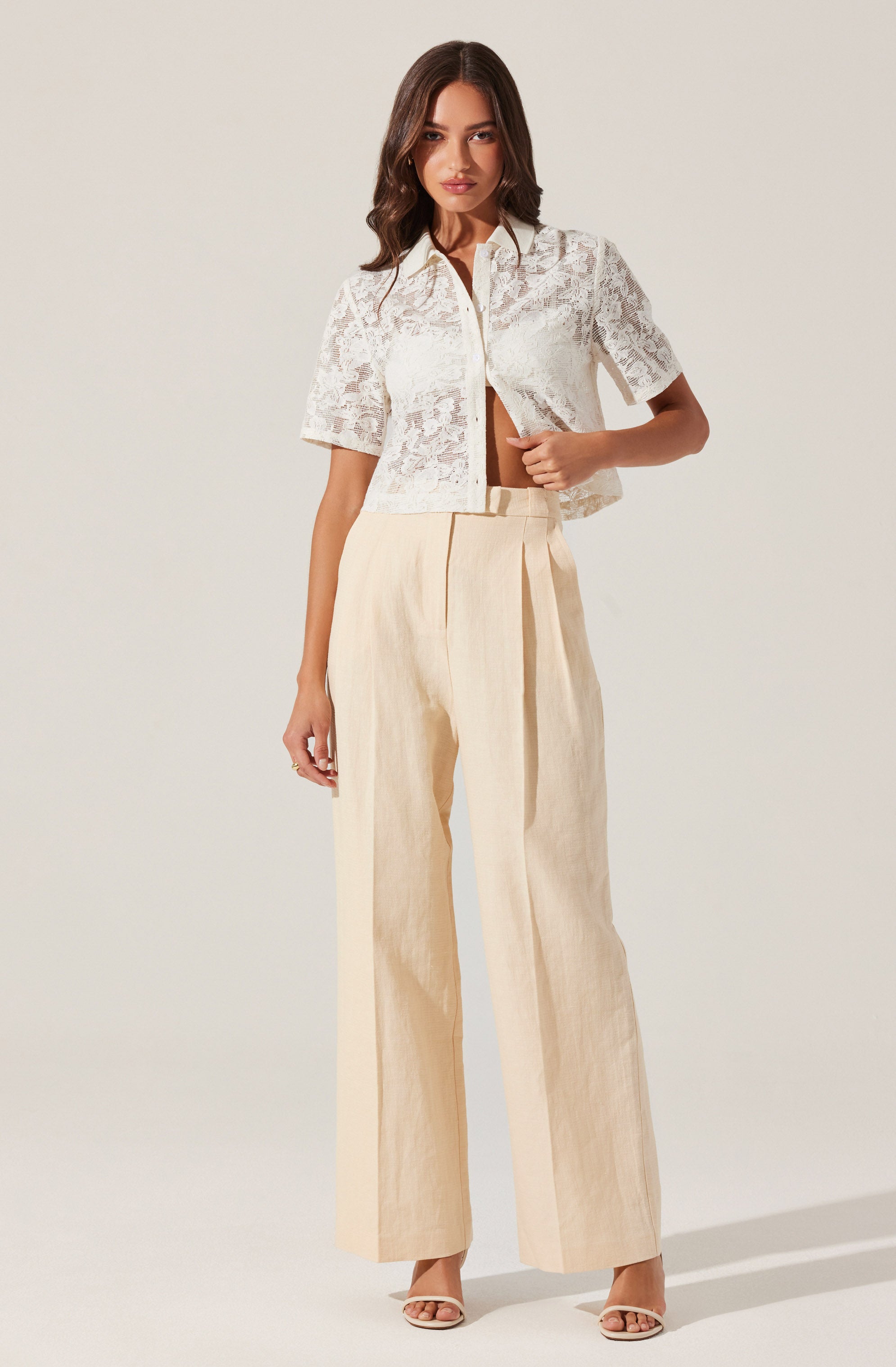 A woman is wearing a short-sleeved, white lace cropped jacket with a collar and front zipper, paired with high-waisted, wide-leg cream trousers. She is also wearing beige open-toed heels.