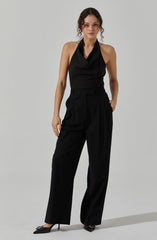 A woman is wearing a sleeveless black top with a draped neckline and subtle pinstripes, paired with high-waisted, wide-leg black trousers. She is also wearing black pointed-toe high heels with embellishments.