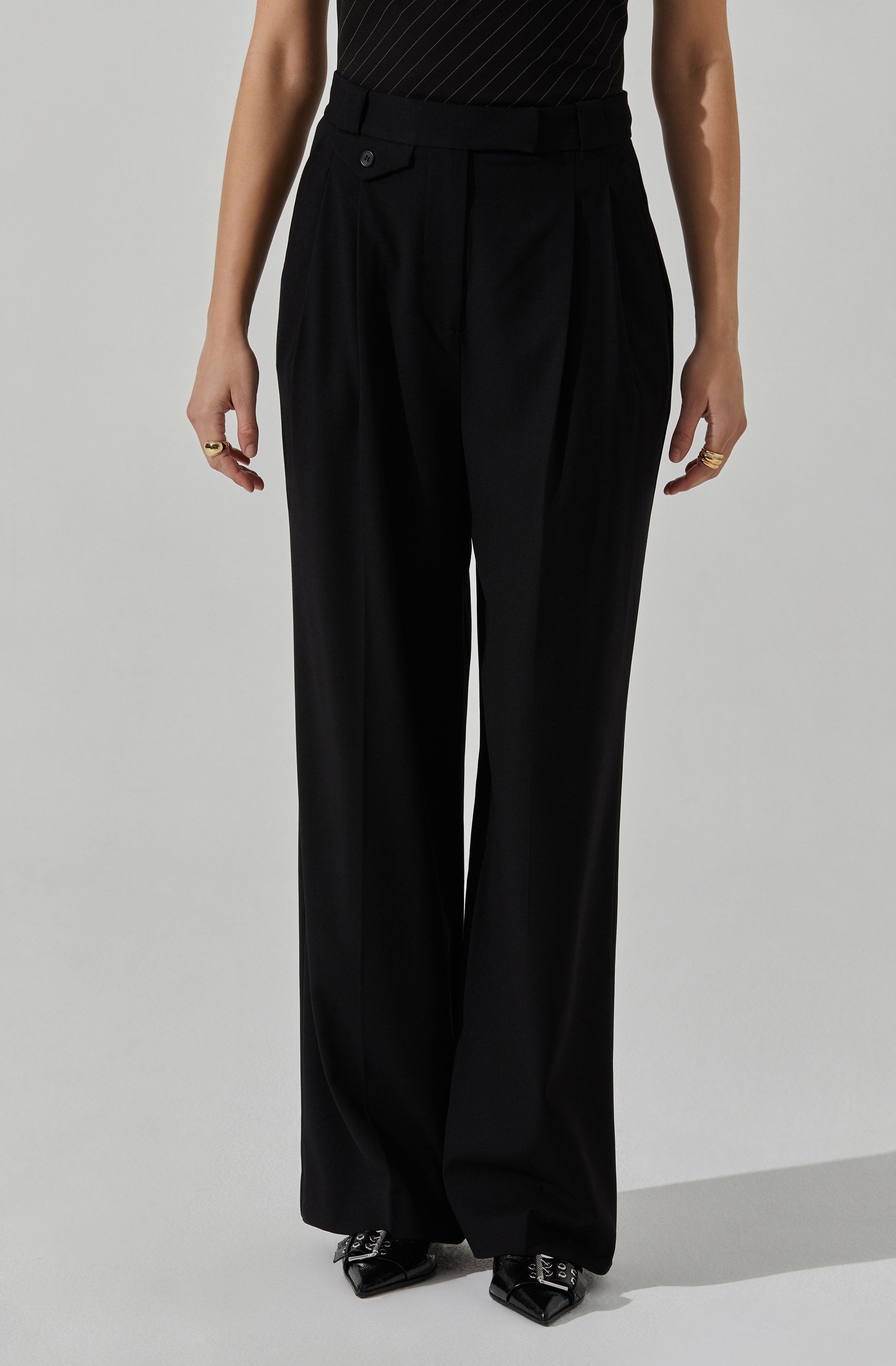 The image shows a person wearing high-waisted, wide-leg black trousers with creases down the front. The trousers have a waistband with a belt loop and a button closure. The fabric appears to be smooth and slightly shiny, giving a formal and elegant look. The person is also wearing a black top with subtle pinstripes. Their hands are relaxed at their sides, adorned with multiple rings. They are wearing black pointed shoes with decorative metallic details.
