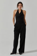 The woman is wearing a sleeveless black halter top with subtle pinstripes and high-waisted, wide-leg black trousers. She has her hands in her pockets and is standing against a plain light-colored background.