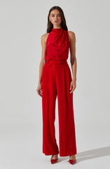 A woman is wearing a sleeveless, high-neck red top made of shiny, silky fabric with a draped front. She is also dressed in high-waisted, wide-leg red trousers with pleats, creating a smooth, flowing silhouette. She is wearing matching red pointed-toe heels, and has long, dark hair and gold earrings.