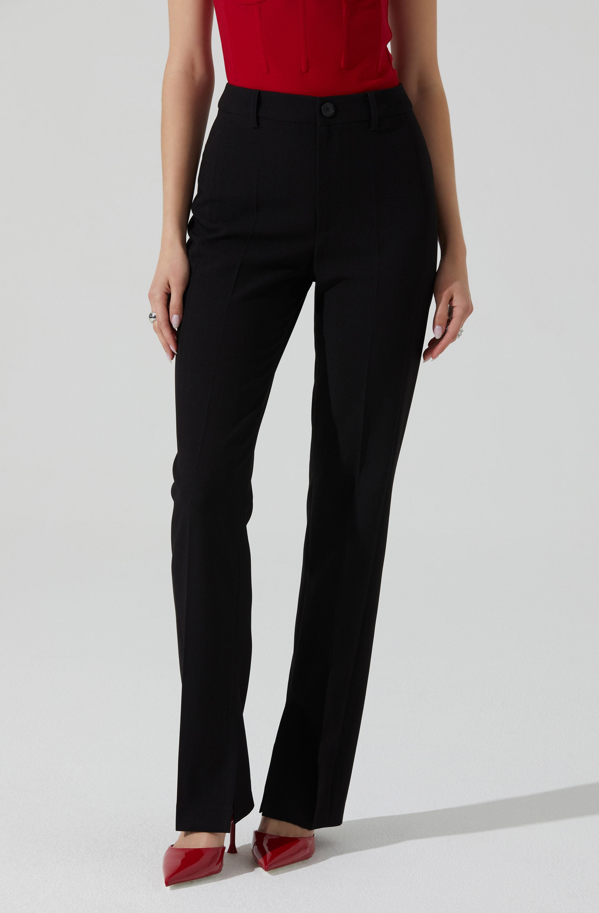 A person is wearing high-waisted black dress pants with a straight-leg cut, secured with a button and zipper at the waist. They are also wearing a sleeveless red top tucked into the pants. The outfit is paired with red pointed-toe high heels.
