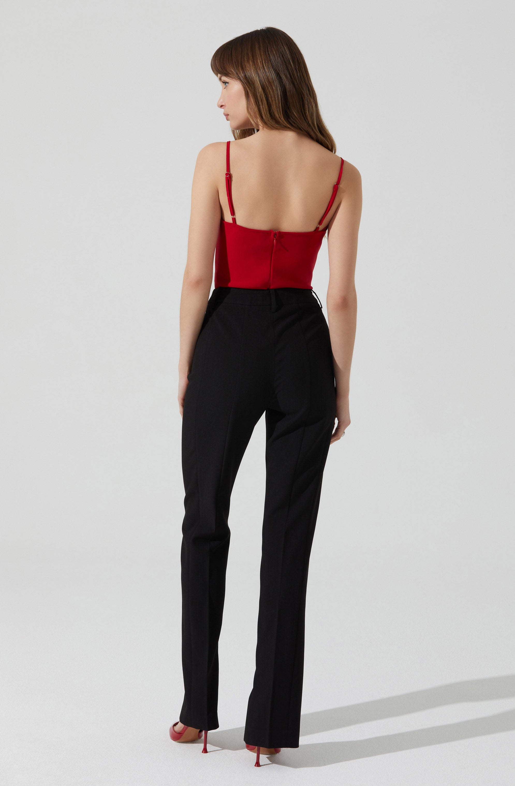 A woman is wearing a fitted red spaghetti strap top with adjustable straps and a zipper closure at the back. She has on high-waisted black trousers that are tailored and slightly loose-fitting, with a crease running down the front of each leg. She is also wearing red high-heeled shoes with thin heels.