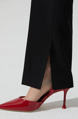 A person is wearing a black pant with a slit at the bottom, paired with a shiny red high-heeled shoe that has a pointed toe and a stiletto heel.