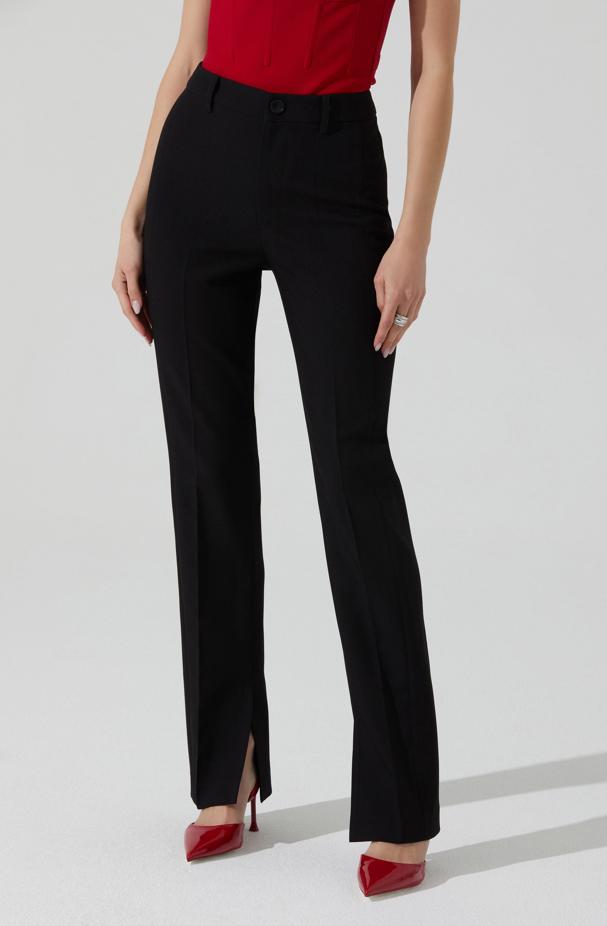 The image shows a person wearing black dress pants with a front button closure and belt loops. The pants have a sleek, tailored fit with a slight flare at the bottom, featuring a small slit at the hem. The person is also wearing red high-heeled shoes.