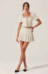 A woman is wearing a cream-colored, textured two-piece outfit consisting of a short-sleeved, cropped jacket with a rounded neckline and front button closure, and a matching pleated mini skirt. The fabric has a woven pattern with subtle checks and a decorative trim along the edges of the jacket and neckline.