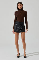 A woman is wearing a fitted, long-sleeve, brown top with a high neckline and cut-out details near the collarbone and shoulders. She pairs it with a black leather mini skirt featuring a gathered, ruched design on one side. She completes the outfit with black pointed high heels that have a small bow detail.