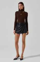 A woman is wearing a fitted long-sleeve brown top with a high neckline and cut-out details near the collarbone. She pairs it with a black, shiny, faux leather mini skirt that has a ruched, gathered design on one side. She completes her outfit with black pointed-toe high heels that have bows on the front.