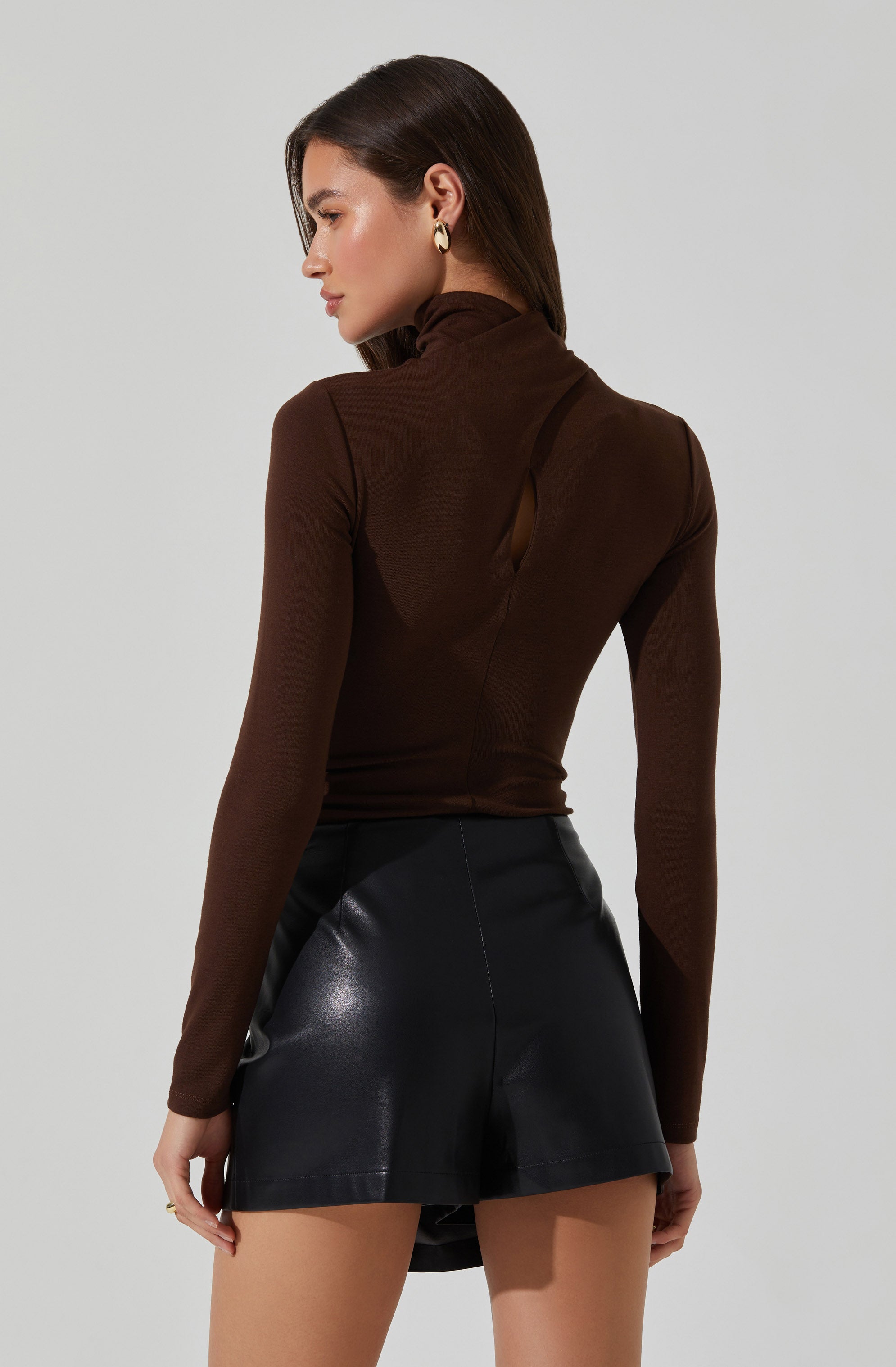 A woman is wearing a fitted, long-sleeved, brown turtleneck top with a small keyhole cutout at the back. She pairs it with high-waisted black leather shorts.