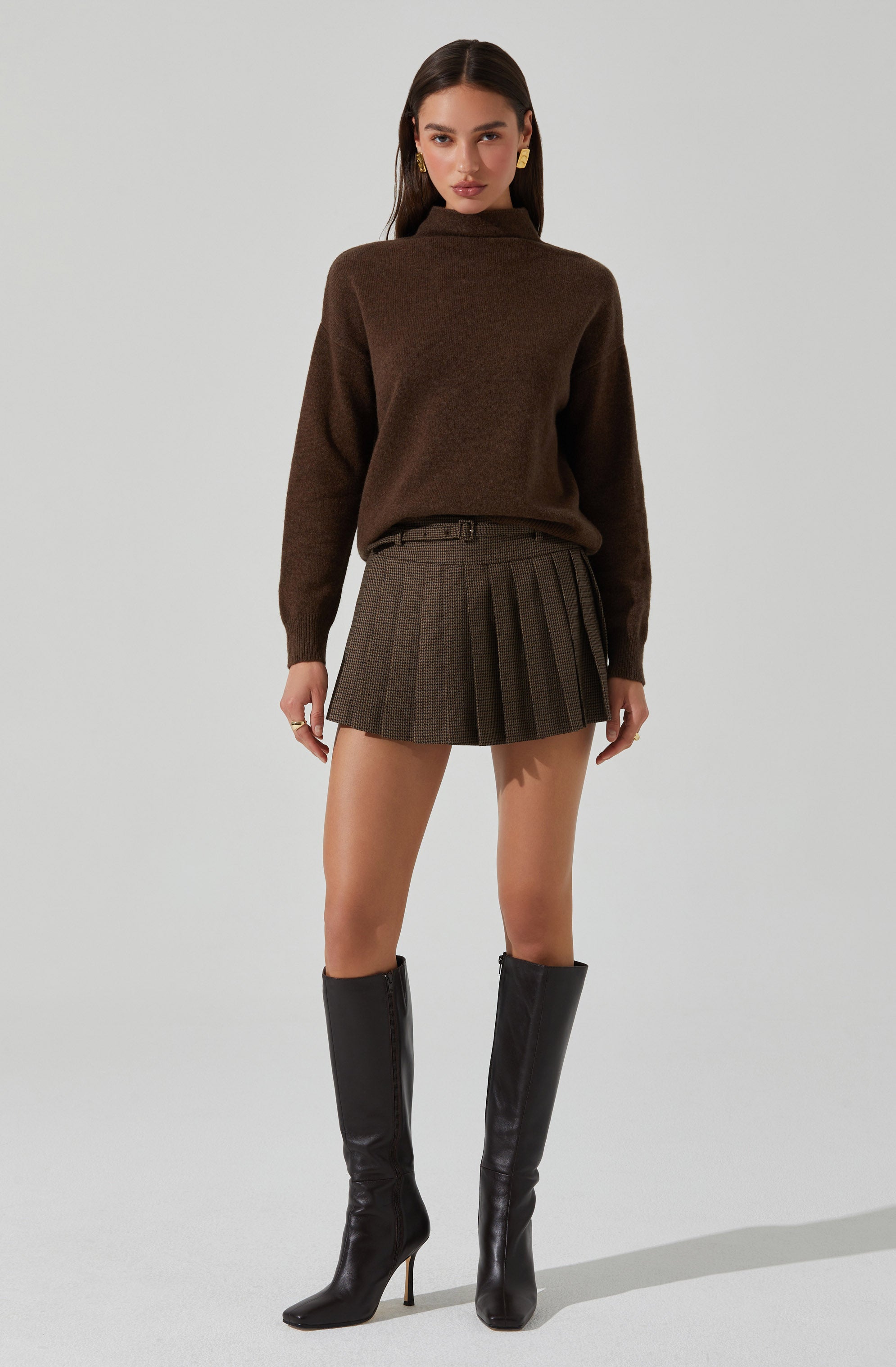 A woman is wearing a brown high-neck sweater with long sleeves, a belt, a plaid pleated skirt with a mix of brown and gray tones, and black knee-high heeled boots. She has long dark hair and is accessorized with large gold earrings.