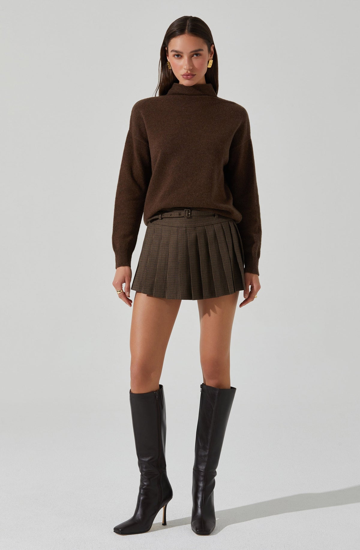 A woman is wearing a brown high-neck sweater with long sleeves, a belt, a plaid pleated skirt with a mix of brown and gray tones, and black knee-high heeled boots. She has long dark hair and is accessorized with large gold earrings.
