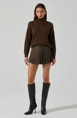 A woman is wearing a brown high-neck sweater with long sleeves, a belt, a plaid pleated skirt with a mix of brown and gray tones, and black knee-high heeled boots. She has long dark hair and is accessorized with large gold earrings.