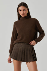 A woman is wearing a dark brown turtleneck sweater with long sleeves, paired with a high-waisted pleated skirt featuring a small checkered pattern in shades of brown and beige. She accessorizes with gold earrings and a ring, with her dark hair parted in the middle and posing with one hand on her hip.