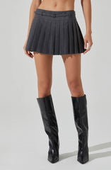 A woman is wearing a high-waisted, dark gray pleated skirt with a belt featuring a small buckle. She is also wearing tall, black, shiny leather boots that reach just below the knee.