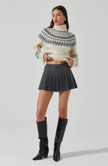 A woman is wearing an oversized cream-colored knit sweater with a high turtleneck and a gray geometric pattern around the shoulders and chest. She pairs it with a high-waisted, dark gray pleated mini skirt with belt loops. She completes the outfit with tall black leather boots that reach just below the knees.