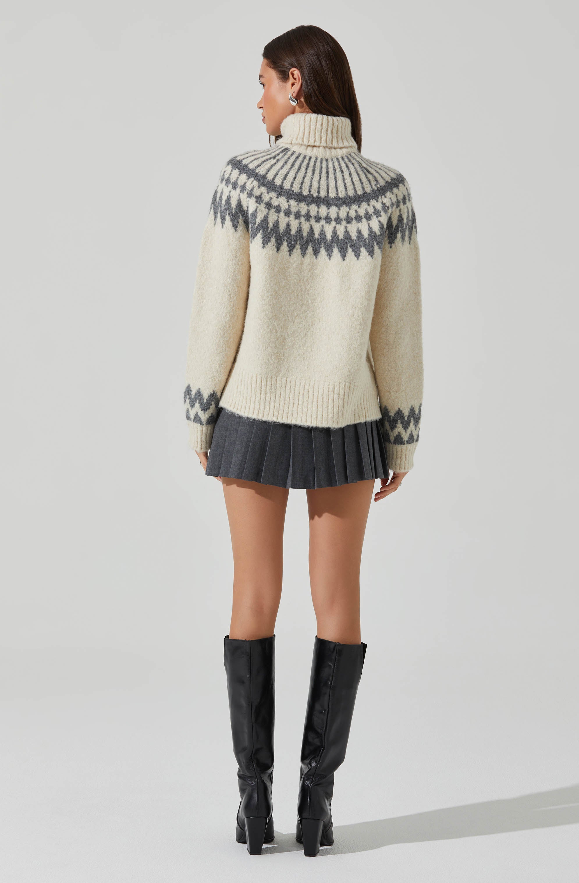 A woman is wearing a cozy, cream-colored turtleneck sweater with a gray geometric pattern around the yoke, sleeves, and cuffs. She pairs it with a short, pleated gray skirt and tall black patent leather boots.
