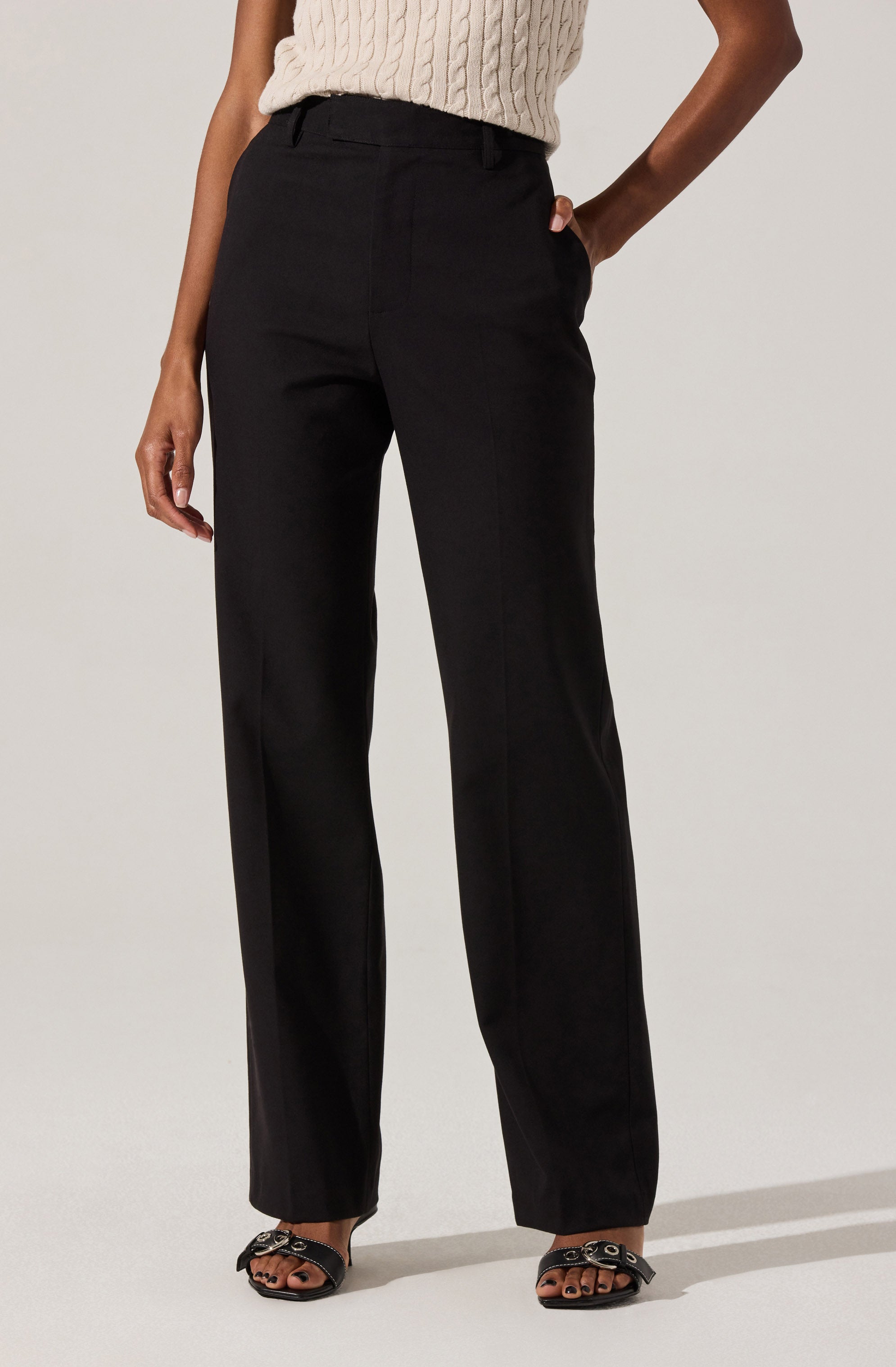 A person is wearing black wide-leg trousers with a high waist and belt loops. They are paired with a cream-colored, knit sleeveless top. The outfit is completed with black open-toe sandals featuring chain details on the straps.