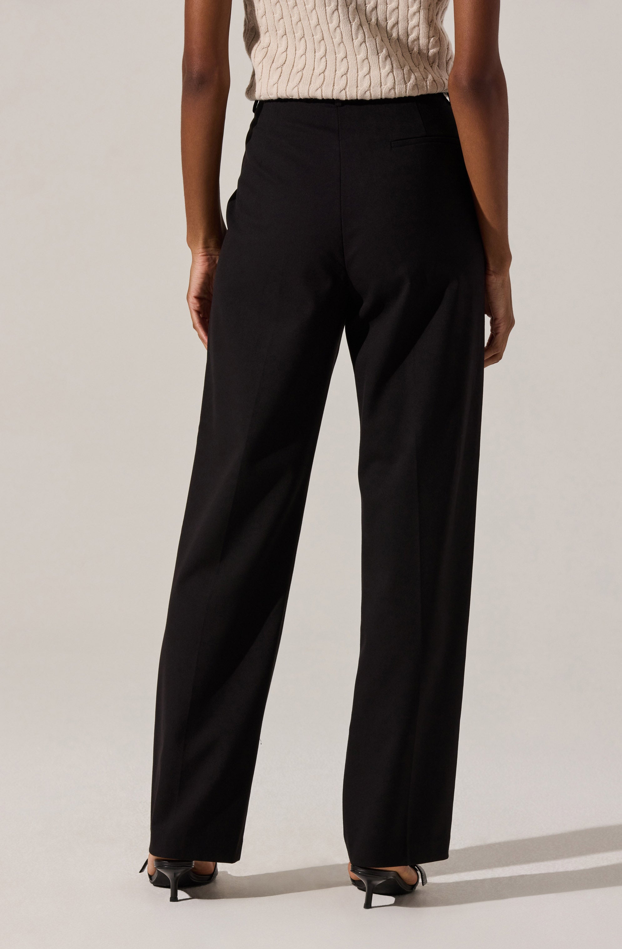 A person wearing high-waisted, wide-leg black trousers paired with a beige, cable-knit sleeveless sweater. They are also wearing black high heels.