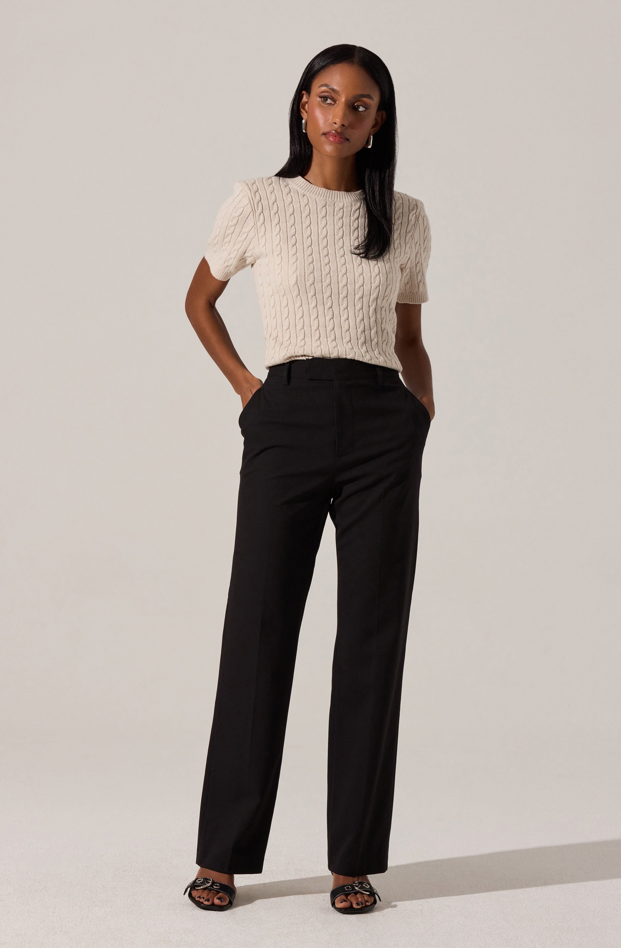 A woman is wearing a cream-colored, short-sleeved cable-knit sweater with a round neckline. She is also wearing high-waisted, straight-leg black trousers and black open-toe heeled sandals with a decorative chain detail.
