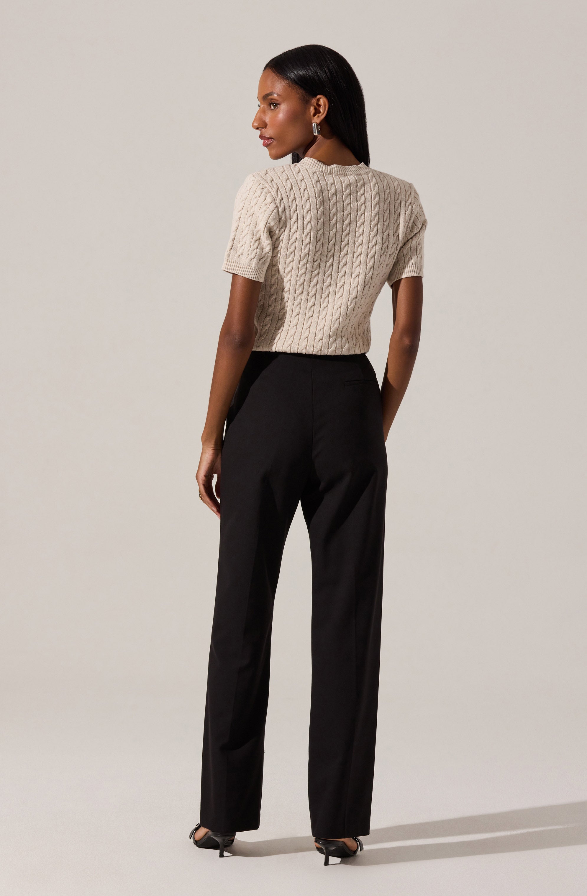 A woman is wearing a beige, short-sleeved, cable-knit sweater and black high-waisted, wide-leg trousers. She has long, straight black hair and is accessorized with earrings and black heels.