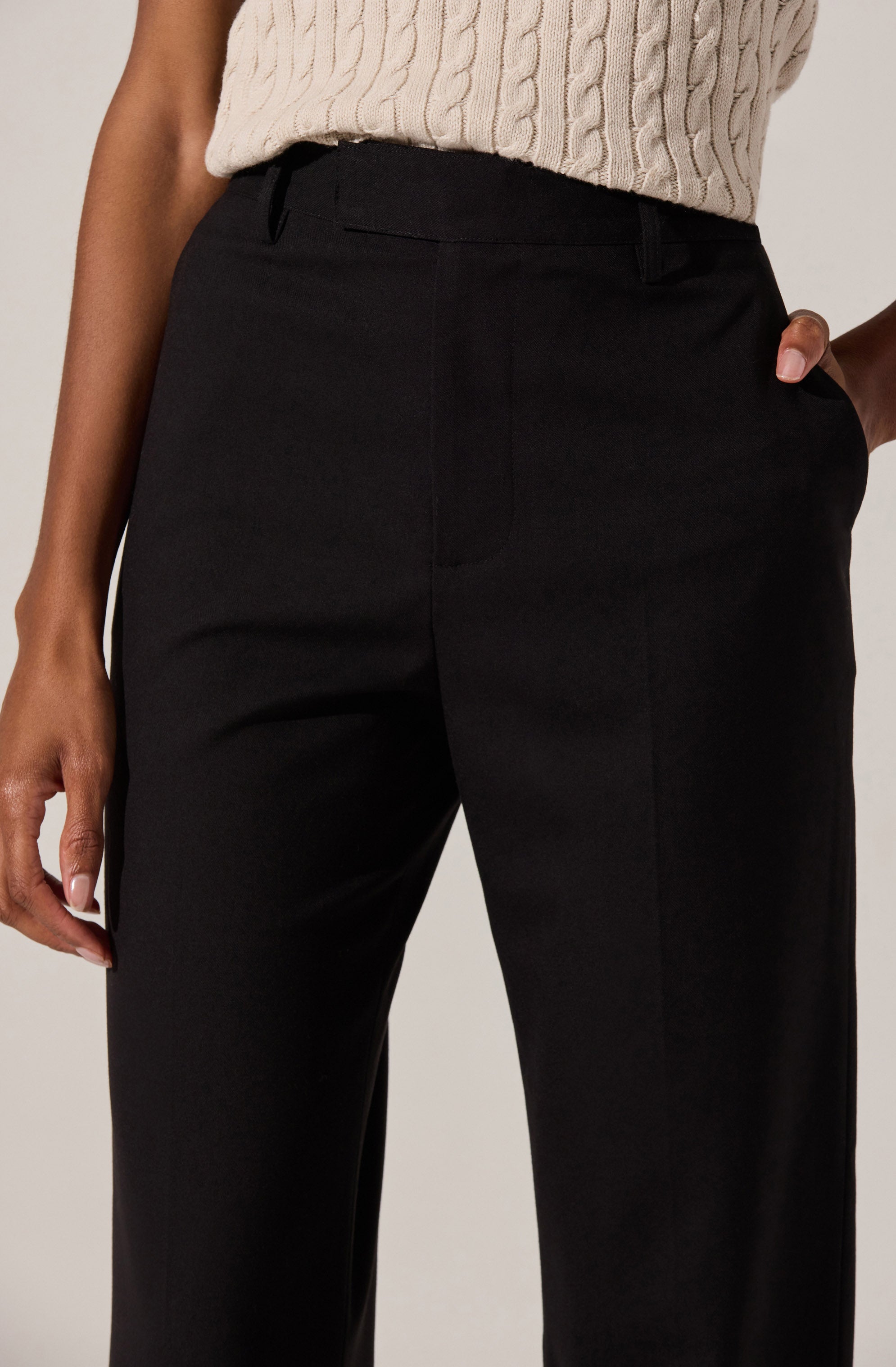 A person is wearing high-waisted black dress pants with belt loops, paired with a cream-colored cable-knit sweater. The pants have a smooth front with no visible pockets, and the person has one hand placed in their pocket.