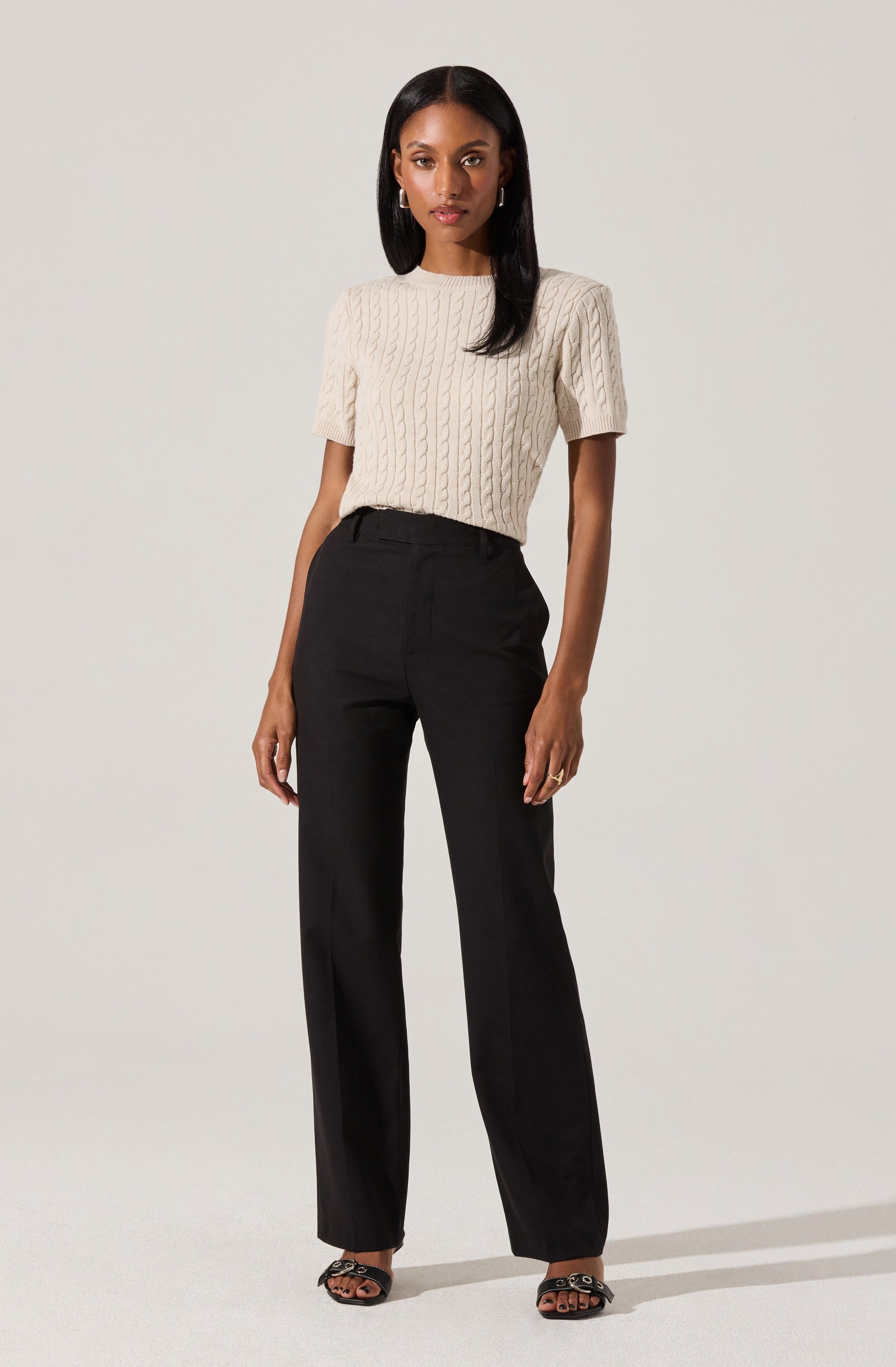 A woman wearing a cream-colored, cable-knit short-sleeve sweater with a round neckline, paired with high-waisted black wide-leg trousers. She is also wearing black open-toe heels with a zipper detail.