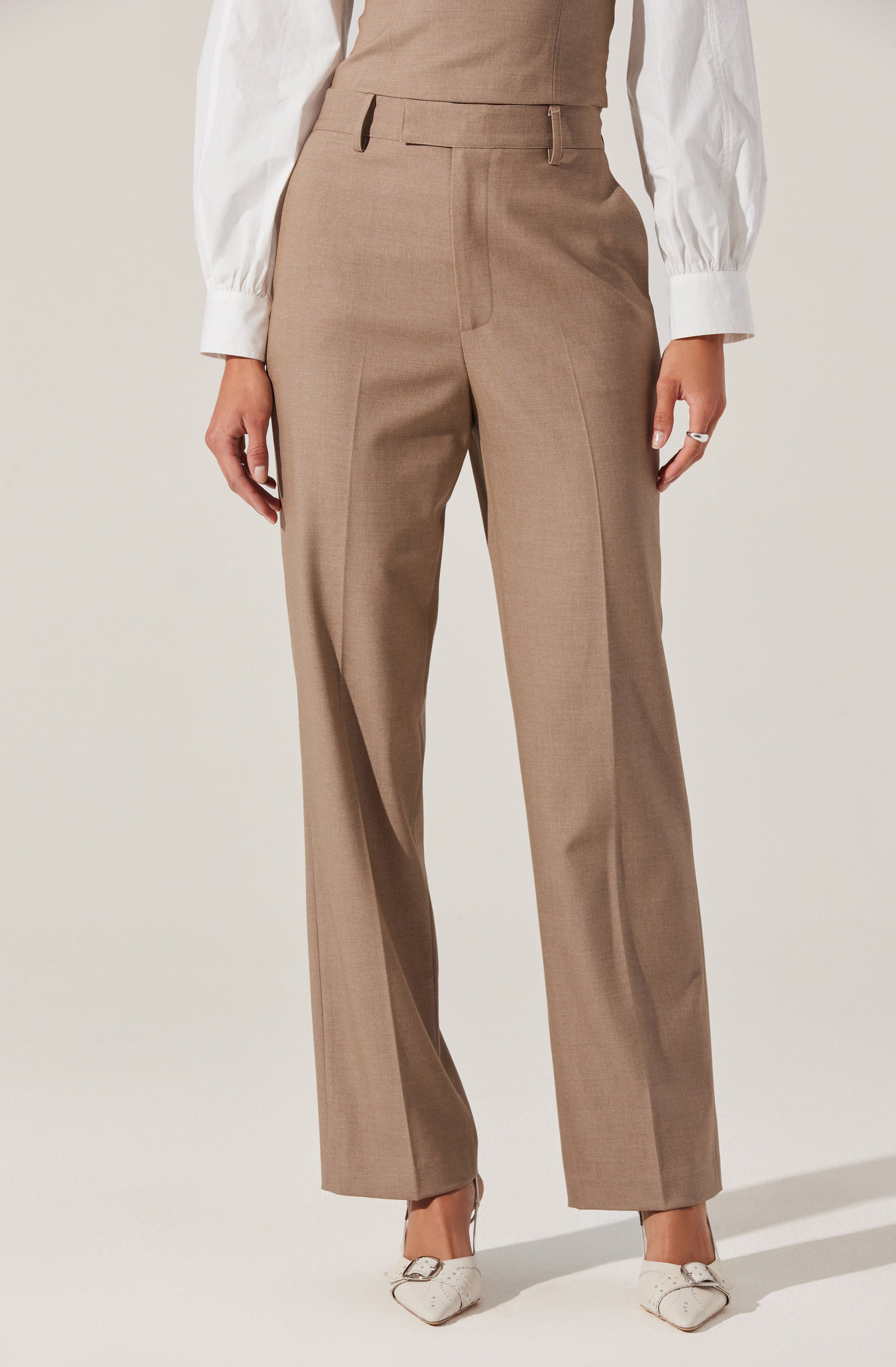 A person is wearing high-waisted, wide-leg tan dress pants with belt loops, paired with a white blouse that has long sleeves with cuffs. The outfit is completed with white pointed-toe shoes.