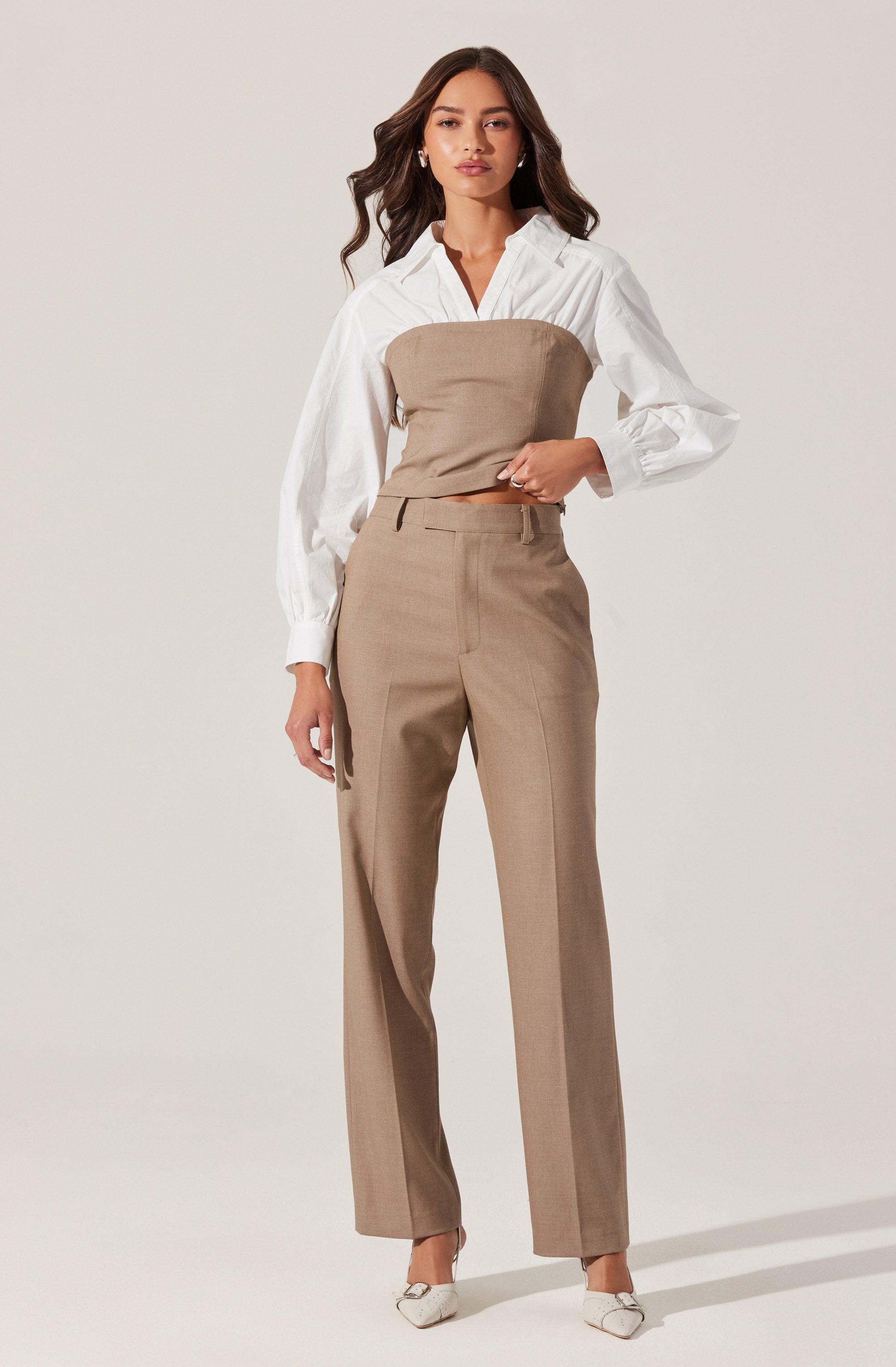 A woman is wearing a white long-sleeve button-up shirt with puffed sleeves, paired with a strapless beige top worn over the shirt. She also has on beige high-waisted, straight-leg trousers with a front crease. She completes the look with white pointed-toe shoes.