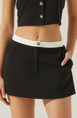 A person is wearing a black cropped sleeveless top with white buttons and a black skirt with a white waistband featuring a single black button. The person’s left hand is resting in the skirt’s pocket.