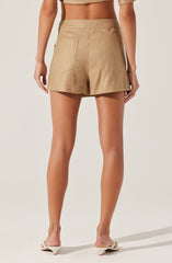The image shows the back view of a person wearing high-waisted beige shorts made of a lightweight fabric, with two patch pockets on the back. The person is also wearing a matching beige crop top and white heeled sandals with a small block heel.