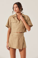 A woman is wearing a beige, short-sleeved, cropped button-up shirt with a collar and two chest pockets, paired with a matching beige high-waisted mini skirt. The shirt has rolled-up sleeves, and the skirt has a front slit and a side button closure.
